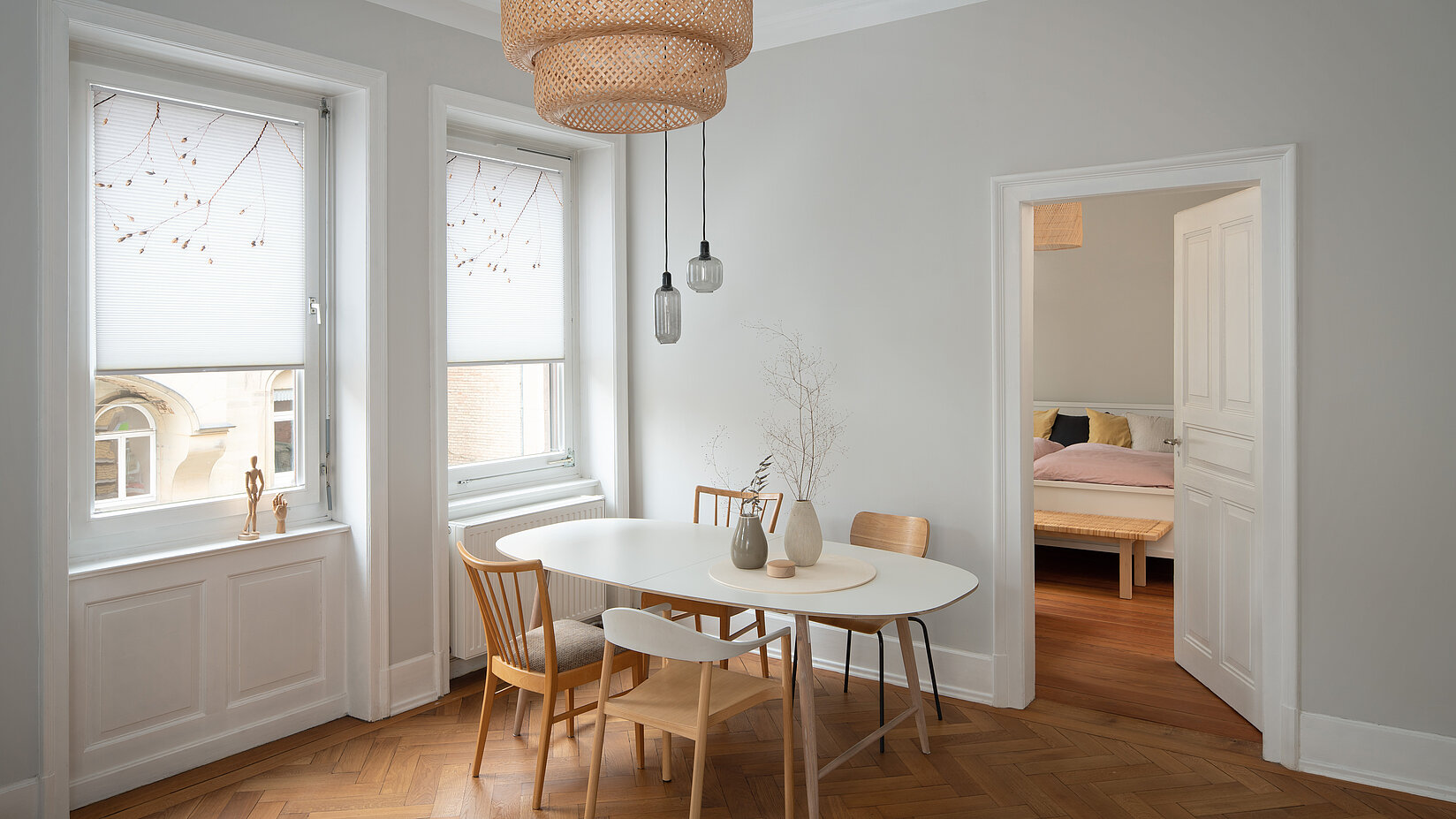 Two tall windows with floral honeycomb blinds in a bright room with parquet flooring and a round dining table.