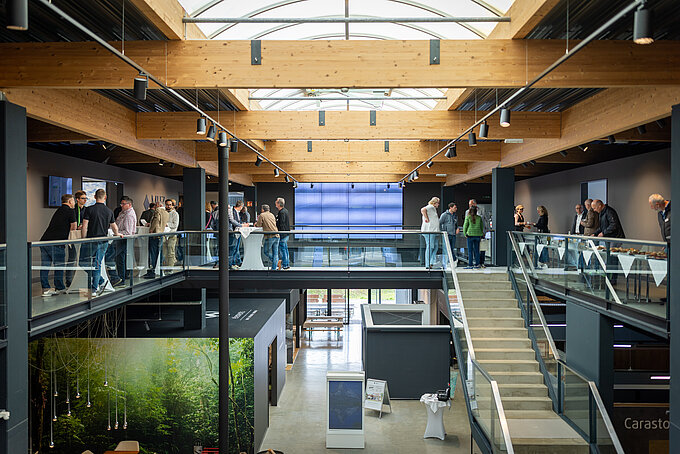 Moderner zweigeschossiger Showroom mit Holzträgern und Glasgeländern, in dem sich Besucher umsehen und unterhalten.