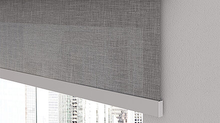 Graues, fein strukturiertes Textilrollo vor einem Fenster mit Blick auf Hochhäuser in einer Großstadt.
