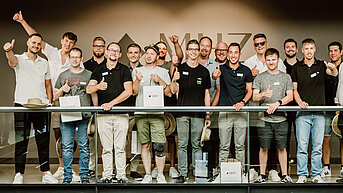 Eine Gruppe junger Männer steht auf einem Balkon und feiert gemeinsam eine Meisterfeier bei MHZ. Einige halten Zertifikate in der Hand, im Hintergrund ist das MHZ-Logo zu sehen.