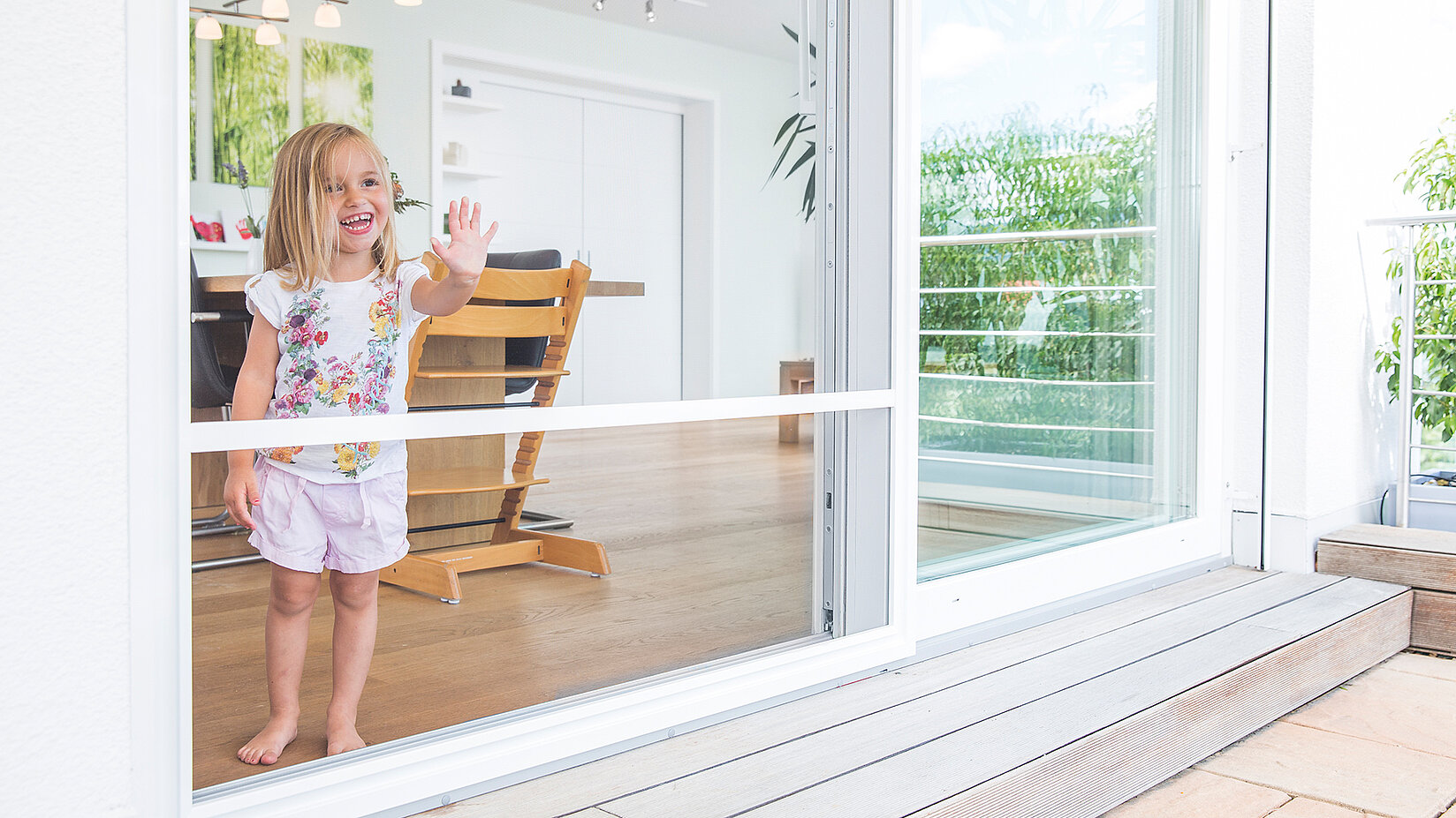 Small girl waves through a closed insect screen sliding door at the patio.