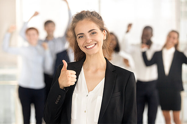 Junge Frau im Business-Outfit zeigt Daumen hoch vor jubelndem Team