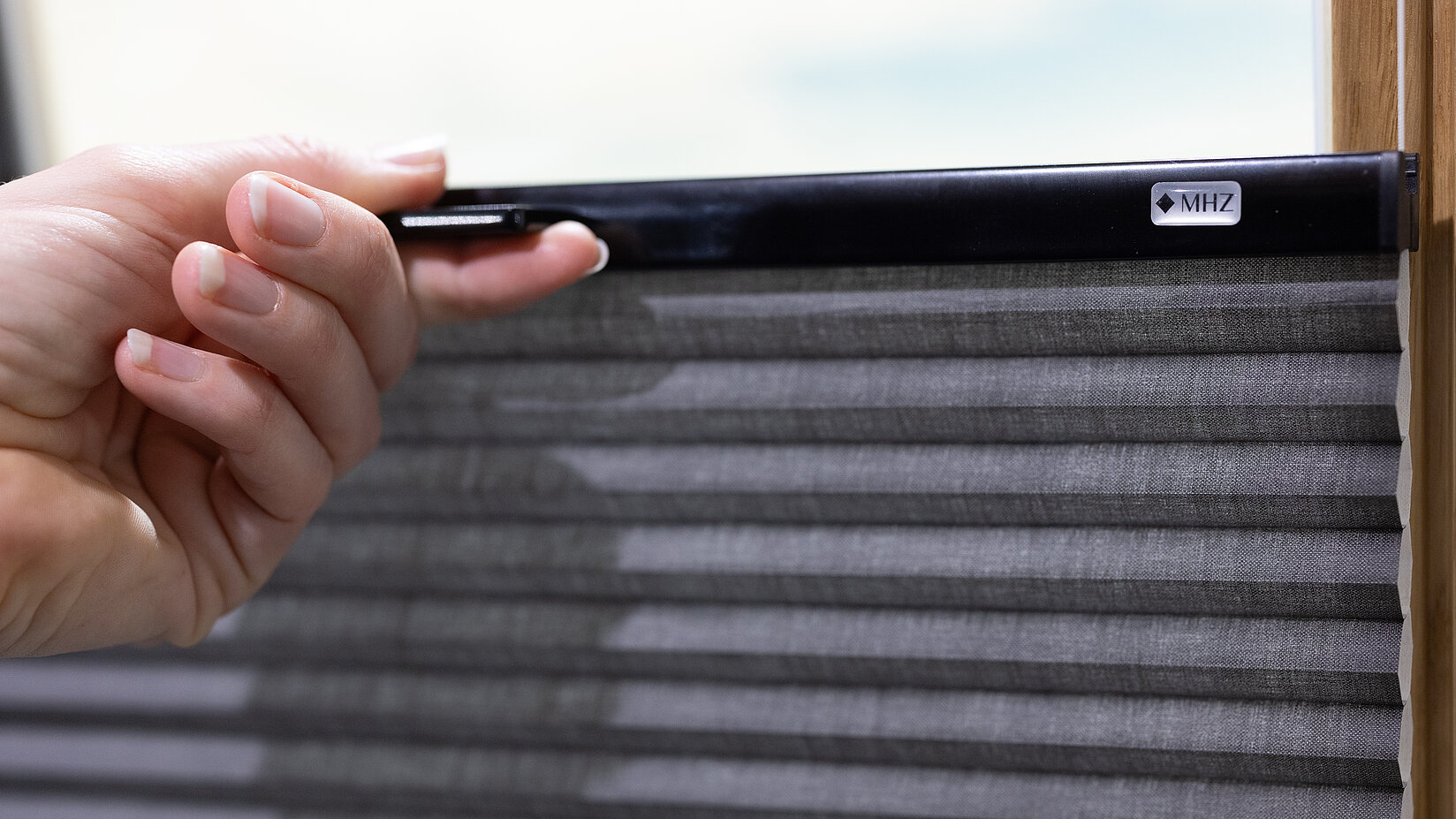 Hand zieht ein dunkelgraues Plissee mit schwarzer MHZ-Bedienleiste an einem Fensterrahmen.
