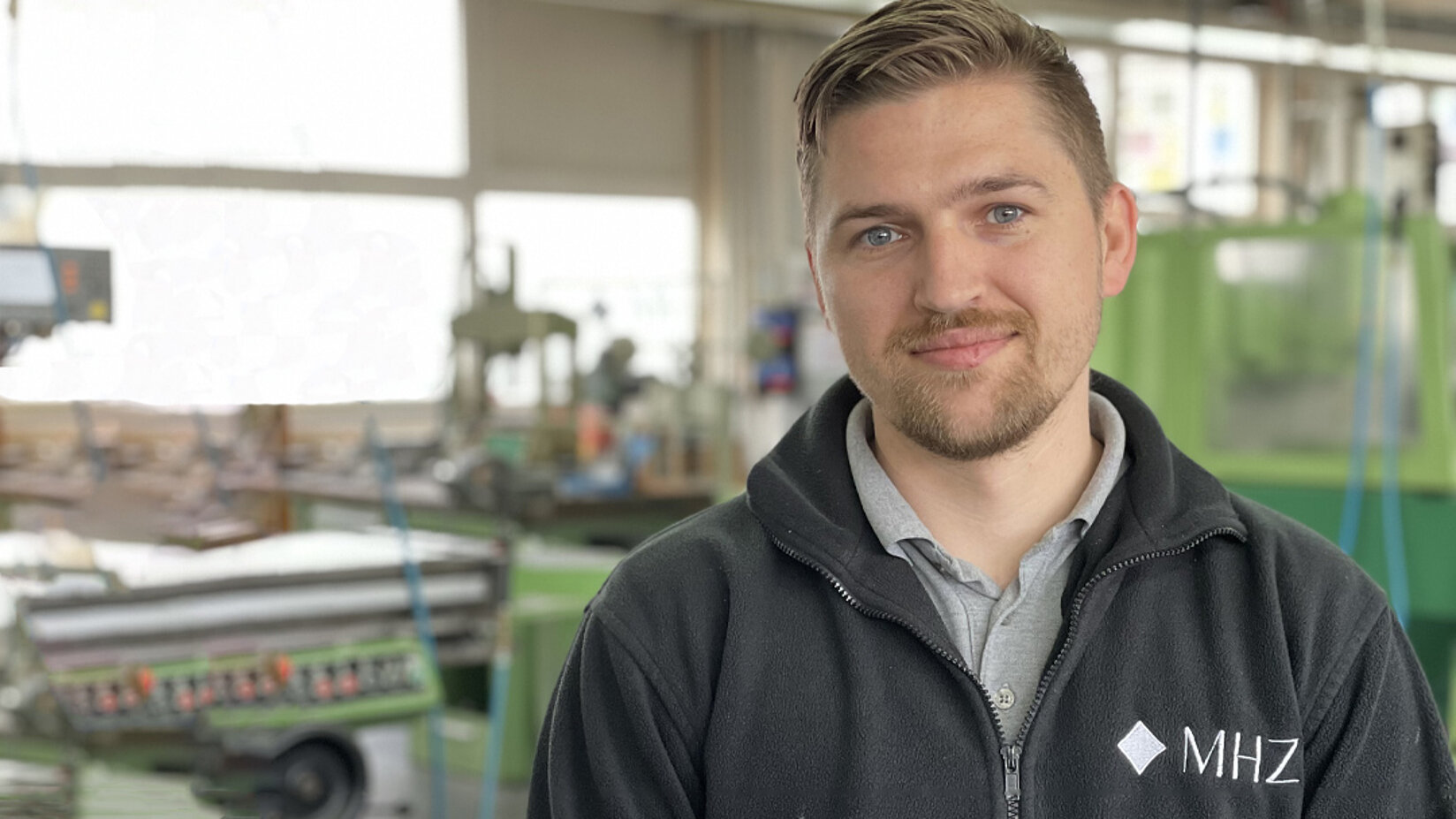Mitarbeiter in Arbeitskleidung mit MHZ-Logo steht in einer Produktionshalle mit Maschinen im Hintergrund.