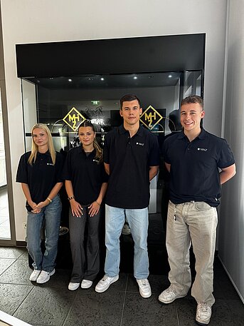 Vier junge Mitarbeitende von MHZ stehen gemeinsam vor einer Glasvitrine und tragen schwarze Poloshirts mit Firmenlogo.