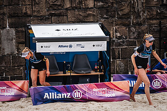 Zwei Beachvolleyball-Spielerinnen vor einer MHZ-Spielerbank bei der German Beach Tour.