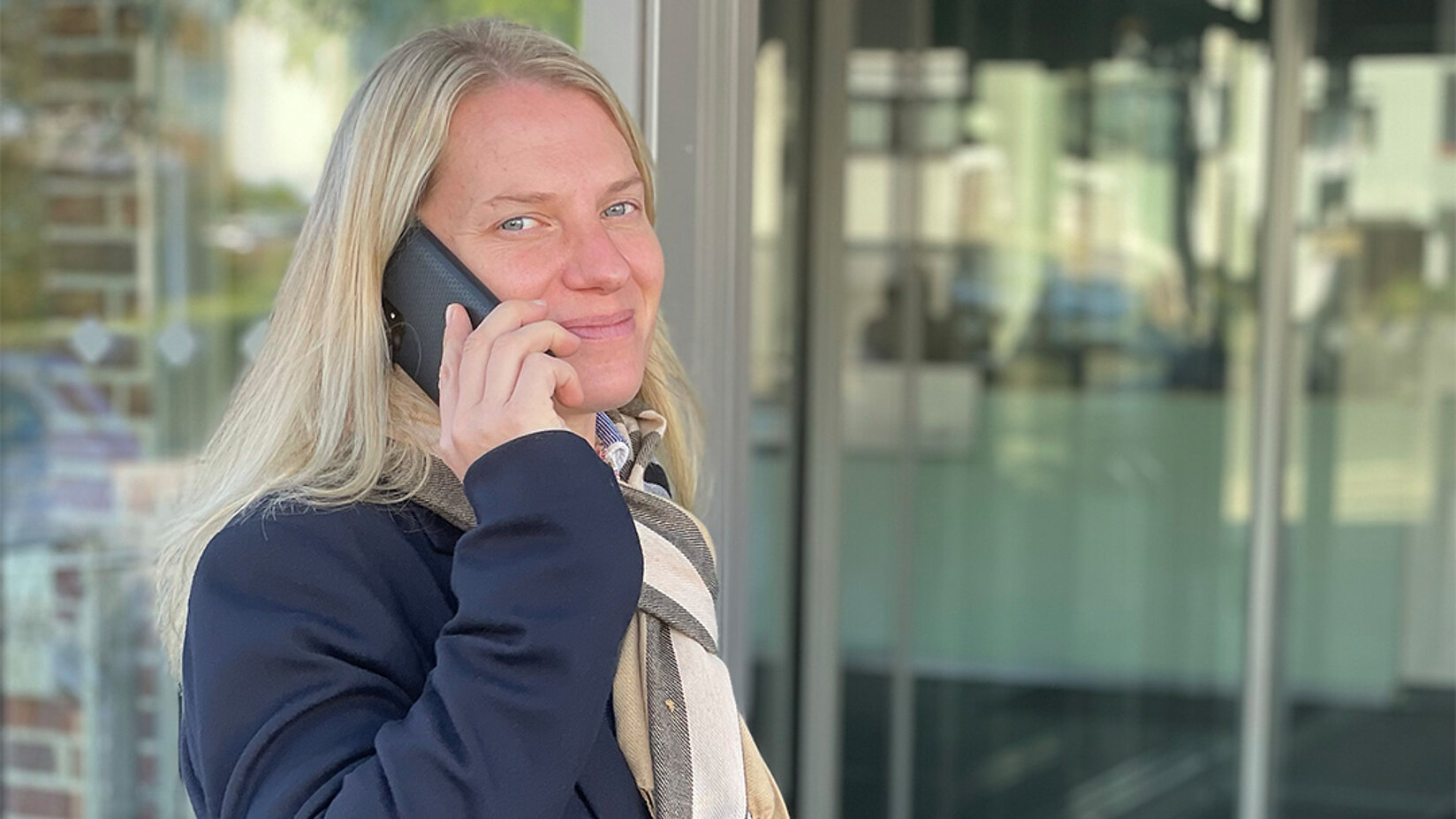 Frau mit blondem Haar telefoniert vor einem Gebäude mit Glasfassade und lächelt.