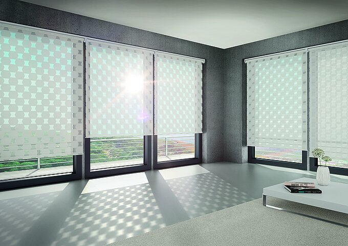 Modernes Wohnzimmer mit großen Fenstern und dem MHZ Doppelrollo „Oscar“ mit grafischem Muster, das dekorative Lichtreflexe auf Boden und Wand wirft.