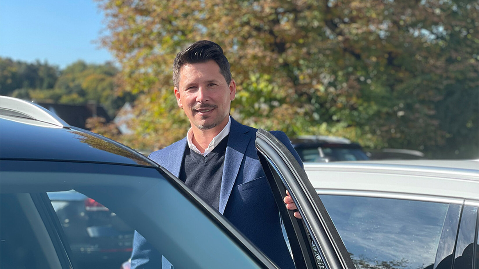 Mann in Businesskleidung steht bei Sonnenschein neben einem Auto auf einem Parkplatz und lächelt.