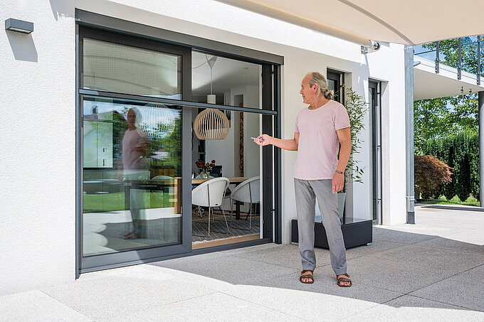 Mann steuert per Fernbedienung ein elektrisches Insektenschutzrollo an einer Terrassentür.