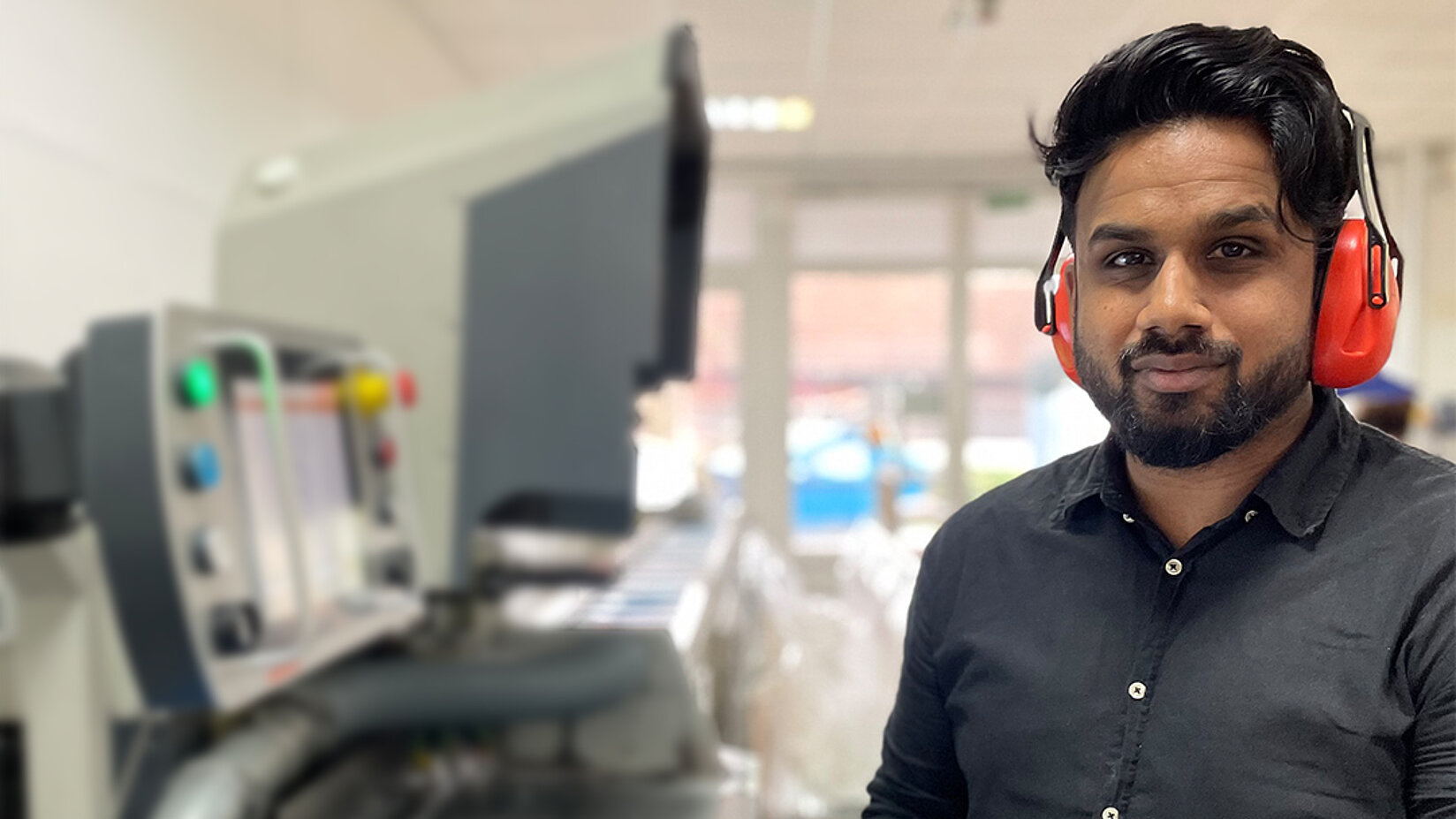 Mann mit Gehörschutz steht neben einer Produktionsmaschine in heller Fertigungshalle.