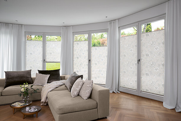 Bright living room with a beige sofa and floral pleated blinds on large windows overlooking a garden.