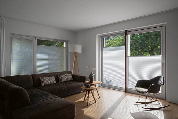 Living room with dark sofa, white pleated blinds on the windows, and a rocking chair in a bright setting.
