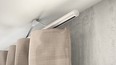 Deckenmontierte Vorhangschiene mit einem beigefarbenen Vorhang in Faltenoptik vor einer Betondecke.