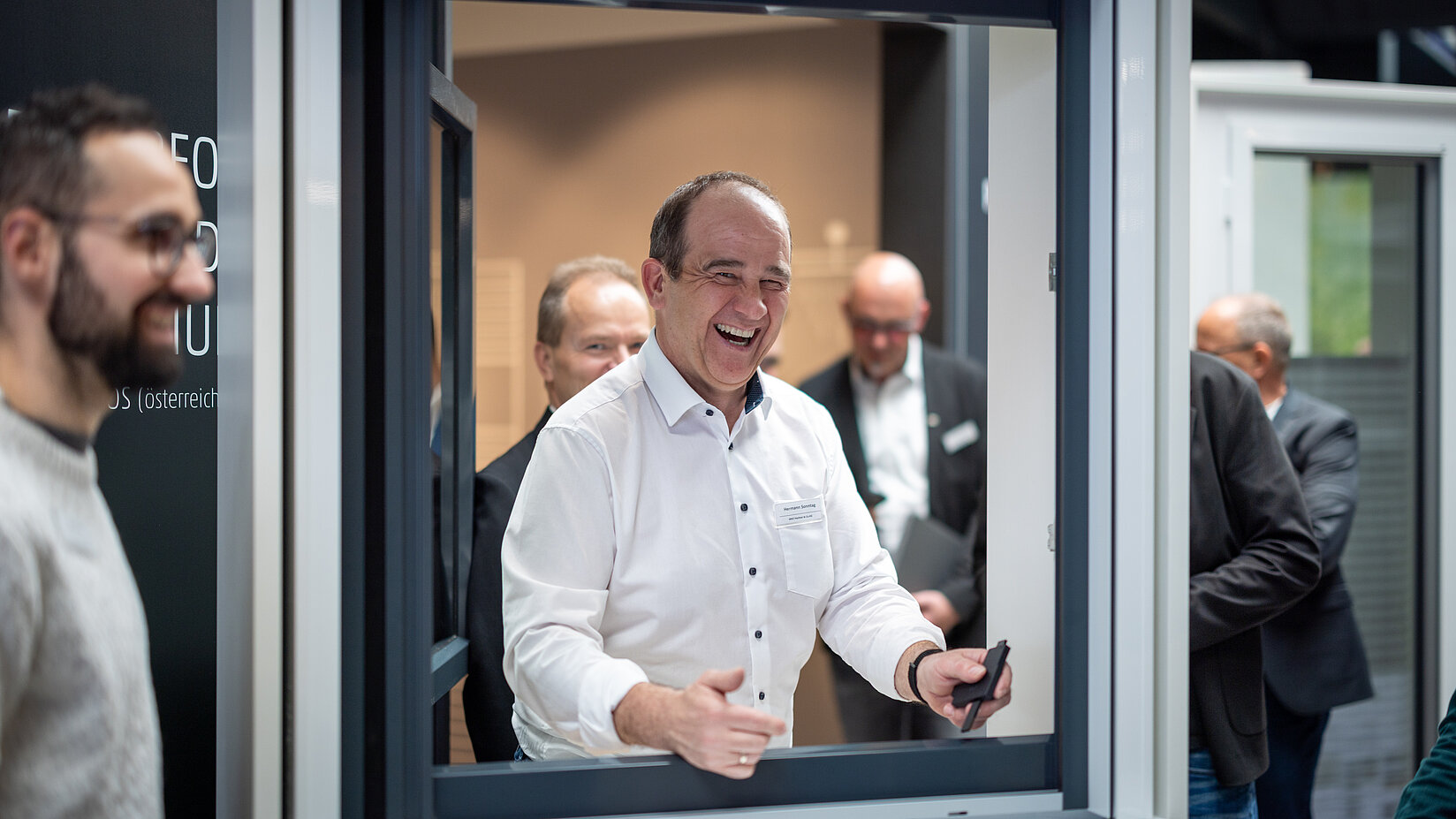 Lächelnder Mann in weißem Hemd steht an einem geöffneten Ausstellungsfenster, umgeben von weiteren Personen in Geschäftskleidung.