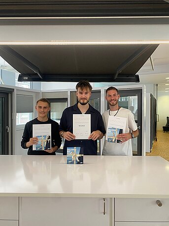 Drei Personen stehen in einer modernen Umgebung und halten Zertifikate in den Händen – sie lächeln stolz in die Kamera.