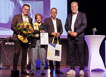 Vier Personen stehen auf einer Bühne. Eine Person hält einen Blumenstrauß und eine Urkunde, daneben stehen zwei Männer mit Geschenktüten und eine weitere Person neben einem weißen Stehtisch. Im Hintergrund ist eine Leinwand mit Projektion zu sehen.