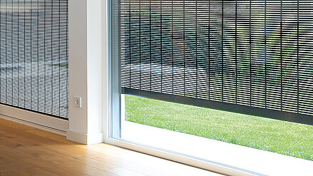 Dunkler, fein strukturierter Metallbehang vor einer Fensterfront mit Blick auf eine grüne Rasenfläche.