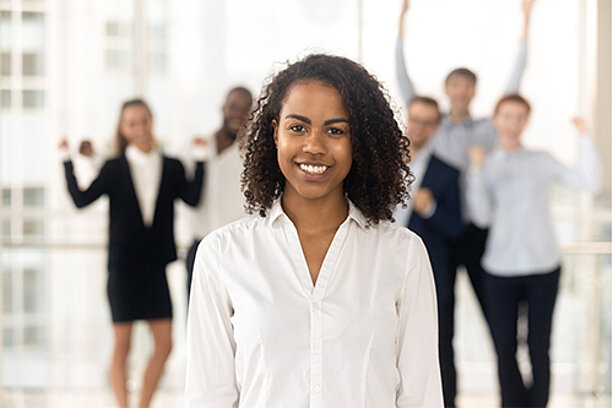 Junge Frau mit lockigem Haar steht lächelnd im Vordergrund, im Hintergrund jubelndes Team vor großen Fensterflächen.