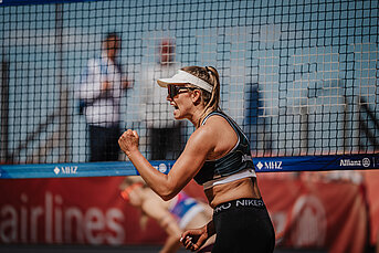 Beach volleyball player cheering with clenched fist in front of net with MHZ branding after winning a point.