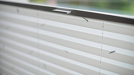 Weißes Plissee mit dezentem Muster vor einem Fenster, mit vertikal verlaufenden Zugschnüren.