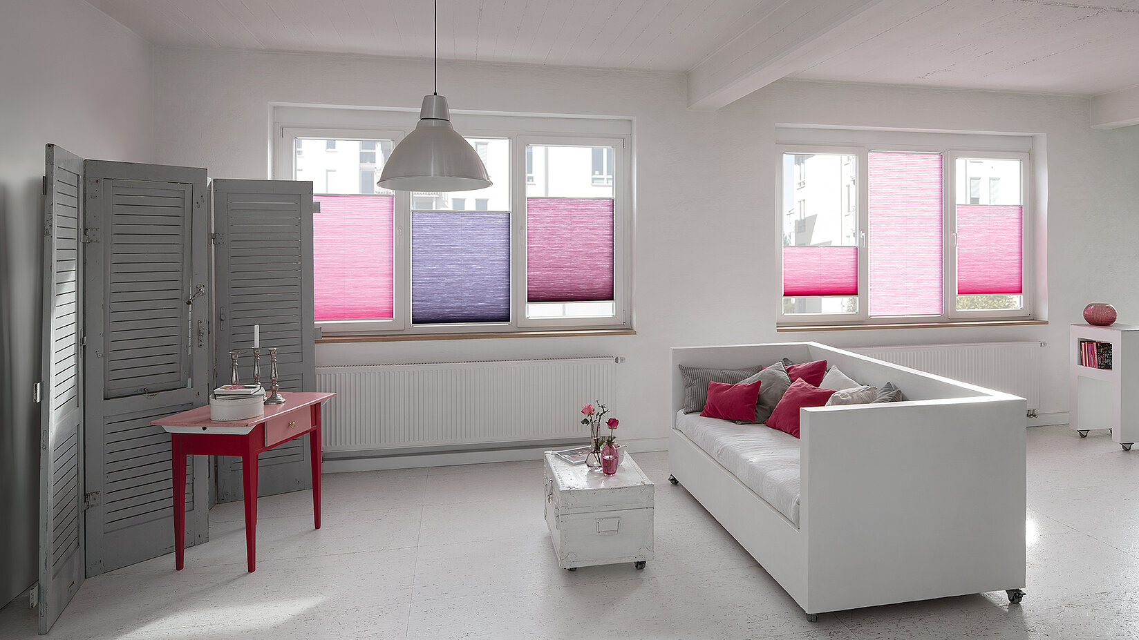 Modernes Wohnzimmer mit farbigen MHZ Duotone-Wabenplissees in Pink- und Violetttönen an weißen Fenstern.