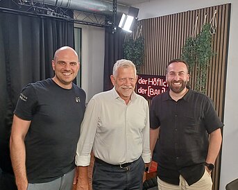 Drei Männer stehen nebeneinander in einem Podcast-Studio und lächeln in die Kamera.